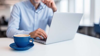 Mann blickt auf Laptop, neben ihm eine blaue Kaffeetasse