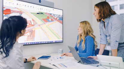 Drei Frauen blicken auf einen großen Bildschirm und beraten über die Skizze einer Autobahntrasse, welche auf diesem zu sehen ist. Auf dem Tisch vor ihnen befinden sich ein Laptop, ein Tablet und einige Dokumente.